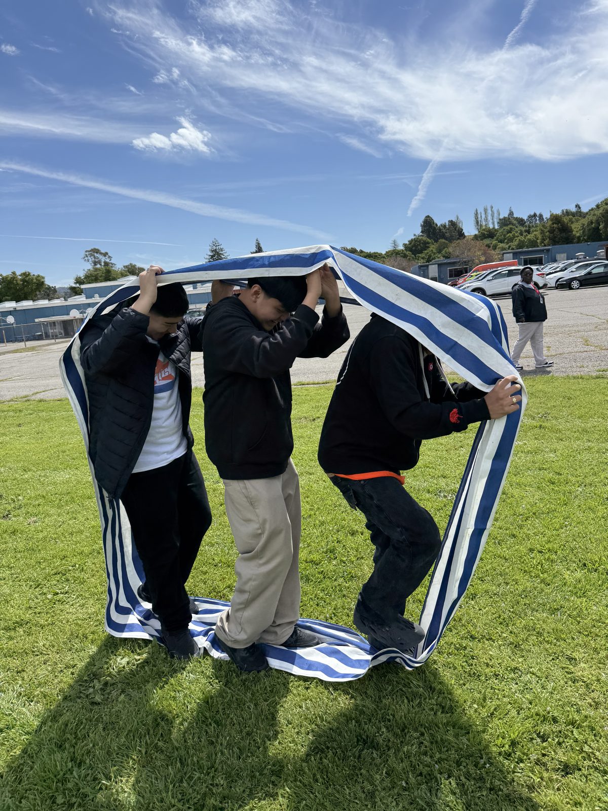 Three students working together outdoors, holding a striped blanket between them
