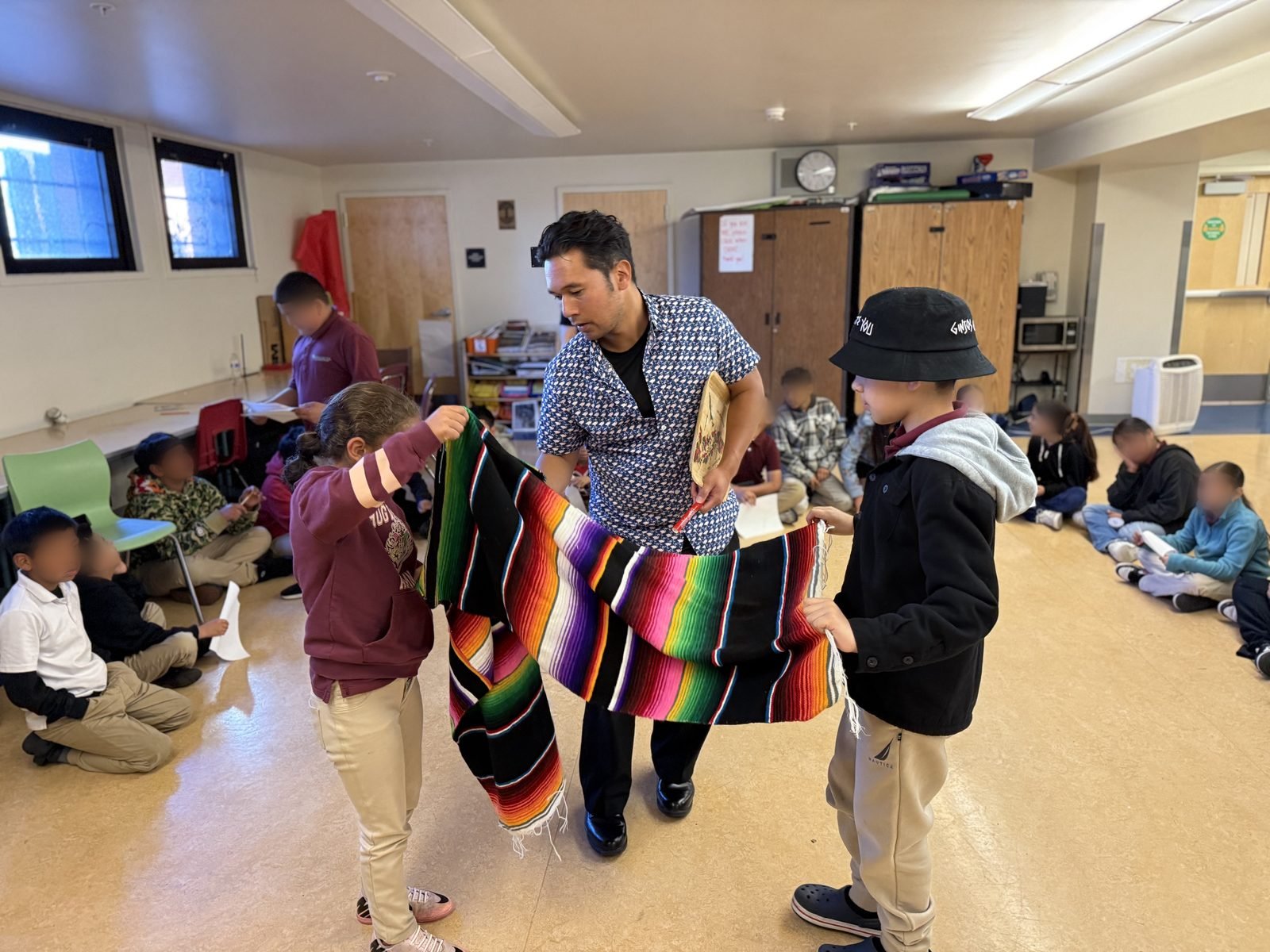 A Zymbolic facilitator leading a classroom activity as two students hold a woven serape